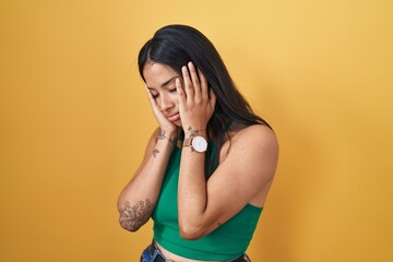 Brunette woman standing over yellow background with sad expression covering face with hands while...