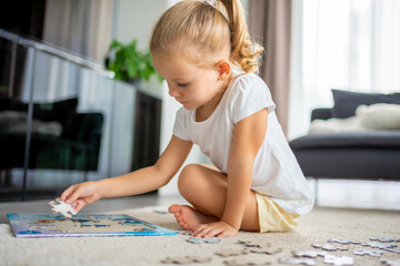 Little blonde girl sits at home on the carpet and collects puzzles