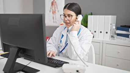 Young hispanic woman doctor using computer speaking on telephone at clinic