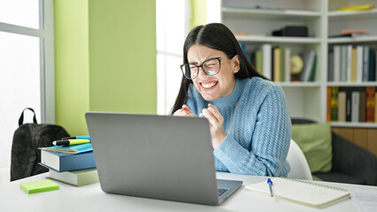 Young hispanic woman student using laptop celebrating at library university