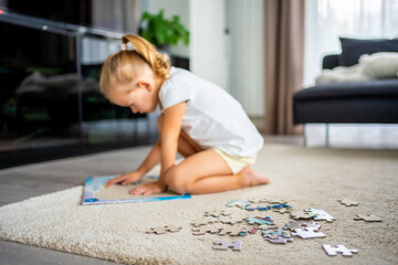 Little blonde girl sits at home on the carpet and collects puzzles