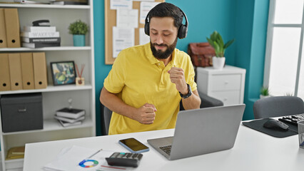 Young hispanic man business worker listening to music dancing at the office