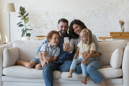 Happy millennial parents holding kids in arms on lap, resting on couch together, taking family home selfie, talking on video call from home, using smartphone, looking at screen, smiling, laughing