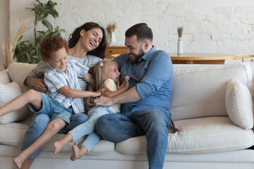 Happy cheerful parents playing with two little kids on sofa at home, enjoying family active leisure time, parenthood, cuddling, tickling giggling children, smiling, having fun - obrazy, fototapety, plakaty
