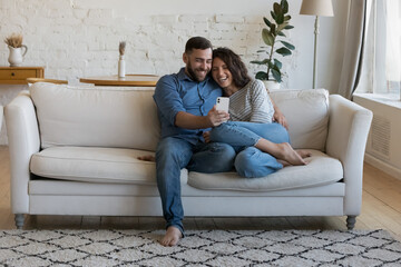 Joyful happy dating couple in love relaxing on home couch, sharing mobile phone, speaking on video call together, enjoying online communication, using Internet app, taking selfie. Full length shot