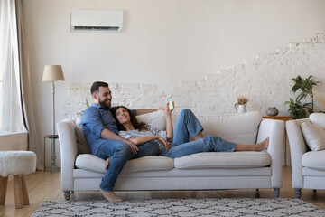 Happy millennial husband and wife resting on couch at home, relaxing under cooling air from...