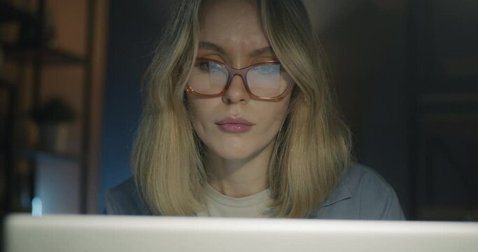 Close-up of pensive businesswoman working with laptop computer looking at screen in dark office in evening. Workplace and technology concept.