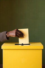 Senior's hand putting yellow vote into ballot box. Unrecognizable person exercising the right to vote.