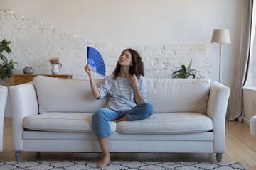 Exhausted frustrated Hispanic woman waving paper handheld fan, cooling hot air, suffering from heat attack, headache, hypoxia, stuffy air, sitting on home couch in living room