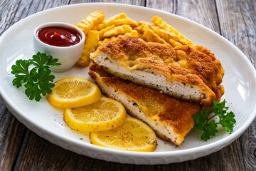 Crispy breaded seared chicken cutlet with French fries, lemon and fresh vegetables on wooden table
