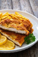 Crispy breaded seared chicken cutlet with French fries, lemon and fresh vegetables on wooden table
