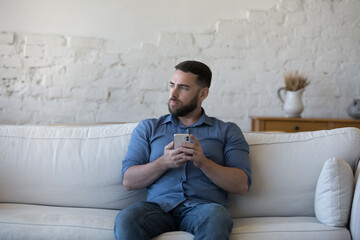 Serious thoughtful mobile phone user man holding gadget, sitting on sofa, looking away, thinking...