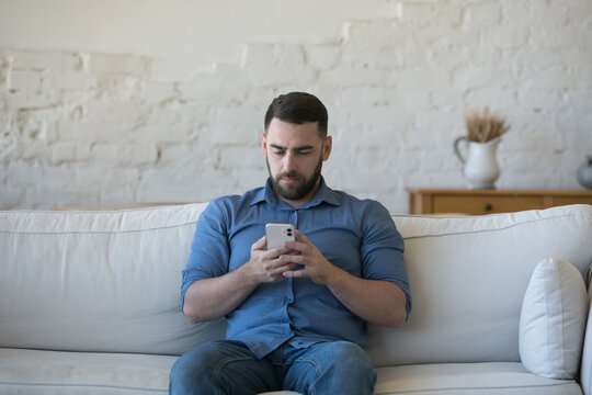 Serious focused millennial man in casual using online virtual app on cellphone at home, sitting on white sofa reading, typing, shopping on Internet, using smartphone for communication