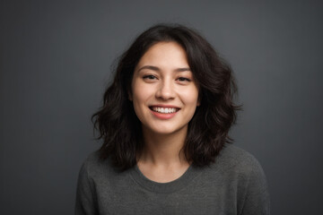 Everyday people. A smiling asian woman with brown wavy hair. Wearing a grey shirt. Pretty woman. On a dark grey background. Healthy glow. Portrait.