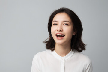 Everyday people. A happy asian woman laughing. Black straight hair to her shoulders. Wearing a white shirt. Pretty woman. University student. On a light grey studio background. Portrait.