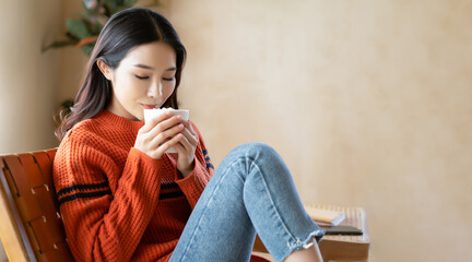 Beautiful Asian woman enjoy a cup of hot cocoa or chocolate with marshmallow while sit on chair in living room at home. Korea girl wearing sweater in an autumn day. Horizontal photo banner for website