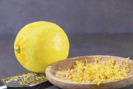 Citron jaune sur une r&acirc;pe en acier, ainsi qu'une assiette en bois contenant les zestes du citron retir&eacute;s &agrave; l'&eacute;corce de l'agrume.