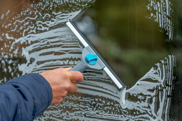 Closeup a hand holding a squeegee tool to clean a dirty glass window with detergent foam on the window. - obrazy, fototapety, plakaty