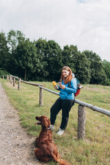 Happy redhead Woman in active trekking clothes with dog having a halt after hiking. Hiker with backpack holding smart phone and drinking water from water bottle or hot drink from yellow thermos