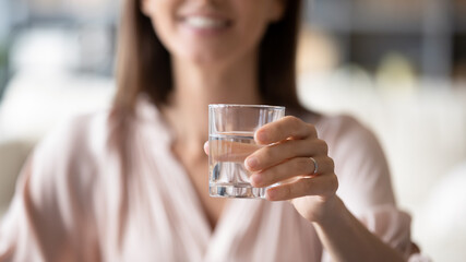 Woman smiles stretched arm close up selective focus of hand holding glass of clean still water, make skin look better, remove toxins from body, remedy, female encourages you healthy lifestyle concept