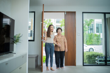 Exiting adult Asian daughter and older mum open the door of new home with smile of happiness, real...