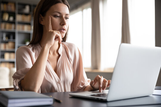 Pensive Woman Sitting At Desk Using Laptop Surfing Website Accesses The Internet Searching Information, Housewife Spend Time On-line, Focused Female Freelancer Do Remote Job At Home Concept