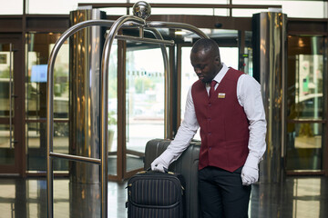 African American doorman putting luggage on trolley standing in lobby of the hotel