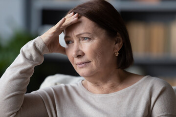 Close up face of attractive 60s woman seated on couch touch forehead with hands thinking about life...