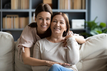 Adult daughter hugs from behind 50s mother seated on sofa, multi-generational female family pose look at camera smiling feels happy. Life value, protection and love, relatives people closeness concept