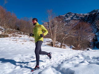 A bearded man is jogging outdoors high in the mountains in winter. A guy in a green jacket and sunglasses is training. Trail running