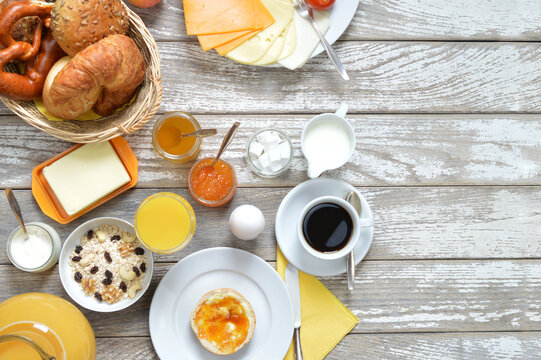 breakfast table with lots of fresh food like coffee, rolls, cheese, sausages, eggs, orange juice, jam, butter and a basket full of croissant and rolls with copy space to the right