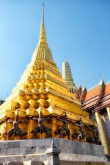 Fototapeta na wymiar A golden temple spire in Thailand, supported by statues of mythical creatures decorated with colorful mosaic tiles, clear blue sky.
