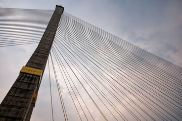 Amidst a cloudy sky, a towering concrete pylon of a cable-stayed bridge commands attention, featuring cables arranged in a harp-like pattern.