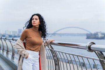 A business woman with black curly hair, lowering her coat from her shoulders, stands upright and...