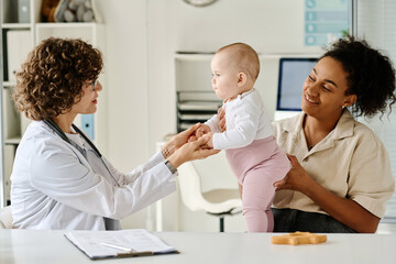 Fototapeta premium Pediatrician examining baby at hospital while mother holding him and listening to doctor
