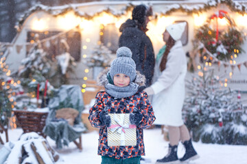 Portrait of a happy child standing on the evening street and holding out hands with gift box at snowy winter day. Christmas gifts, holiday atmosphere.