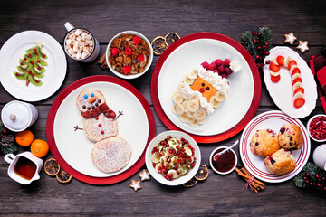 Christmas breakfast table scene. Top down view on a rustic dark wood background. Fun holiday food concept. Santa and snowman pancakes, scones, fruit and cereals.