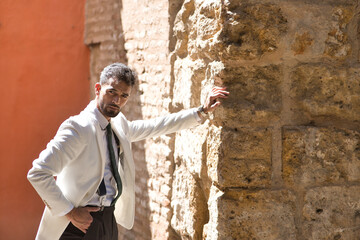 Fototapeta na wymiar Attractive young businessman with beard, suit and tie, posing with hand in pocket leaning against a stone wall looking at camera. Concept beauty, fashion, success, achiever, trend.