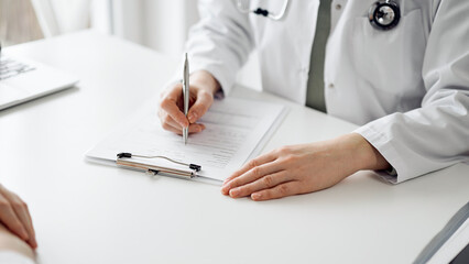 Doctor and patient sitting near each other at the white desk in clinic. Female physician is...