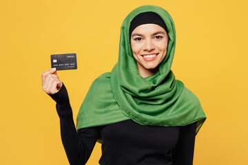 Young arabian asian muslim woman wearing green hijab abaya black clothes hold in hand mock up of credit bank card isolated on plain yellow background People uae middle eastern islam religious concept