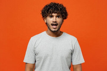 Young sad shocked astonished scared fearful Indian man he wearing t-shirt casual clothes looking camera with opened mouth isolated on orange red color background studio portrait. Lifestyle concept. - obrazy, fototapety, plakaty