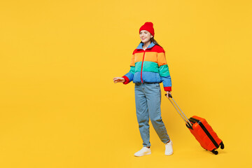 Traveler woman wear padded windbreaker jacket red hat hold suitcase bag go isolated on plain yellow background. Tourist travel abroad in free spare time rest getaway. Air flight trip journey concept.