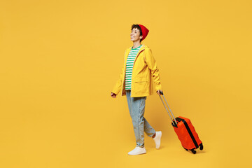 Full body traveler woman wear waterproof raincoat outerwear hold bag suitcase go isolated on plain yellow background. Tourist travel abroad in free time rest getaway Air flight trip journey concept