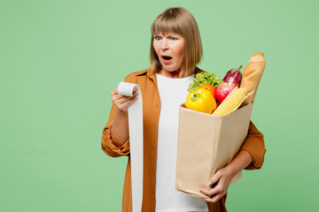 Elderly shocked sad astonished dissatisfied woman wear brown shirt casual clothes hold shopping paper bag with food products read check isolated on plain green background. Delivery service from shop.