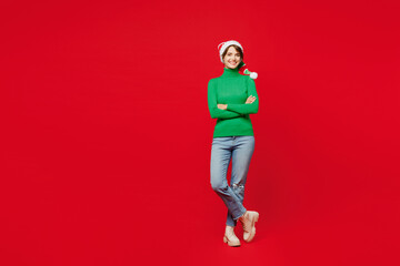 Full body young smiling woman wear green turtleneck Santa hat posing hold hands crossed folded look camera isolated on plain red background. Happy New Year 2024 celebration Christmas holiday concept.