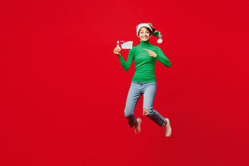 Full body young woman wear green turtleneck Santa hat posing jump high hold store gift coupon voucher card isolated on plain red background. Happy New Year 2024 celebration Christmas holiday concept.