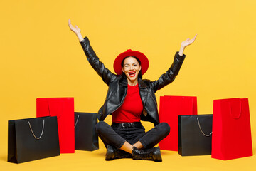 Full body young happy winner woman wearing casual clothes red hat sit near shopping paper package bags spread hands look camera isolated on plain yellow background. Black Friday sale buy day concept.