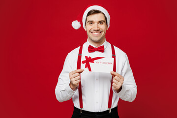 Merry young fun man wear white shirt Santa hat posing hold gift certificate coupon voucher card for...