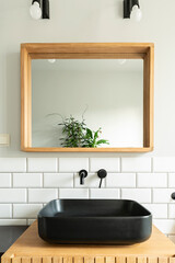 Ceramic black basin on wooden cabinet in industrial bathroom in loft apartment. Stylish interior with square mirror on white tiles on the wall.