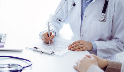 Doctor and patient sitting at the table in clinic. The focus is on female physician's hands filling...